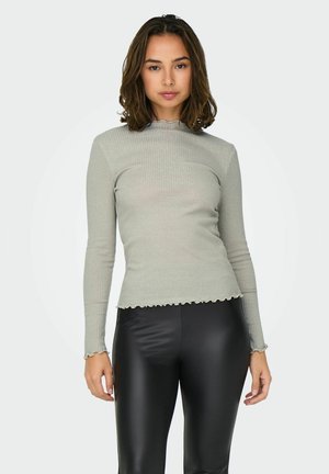 Vrouw met schouderlang bruin haar, gekleed in een lichtgrijze geribbelde lange mouwtop en zwarte leren broek, staand tegen een effen achtergrond.