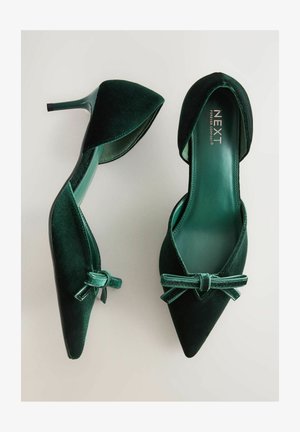 Tamsiai žali aksominiai smailiankulniai bateliai su kulkšnių dirželiais ir mažais kaspinėliais, vienas batelis matomas iš viršaus, kitas šoniniu kampu.