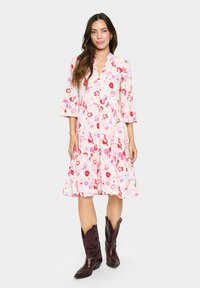 Robe fleurie avec un fond rose, ornée de fleurs rouges et violettes, décolleté en V et manches trois-quarts ; associée à des bottes de cowboy marron foncé.