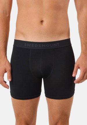 Männlicher Oberkörper, der schwarze Boxer-Shorts mit SWEDEMOUNT-Logo am Bund trägt, vor einem schlichten hellen Hintergrund stehend.