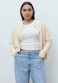 Chaqueta de punto texturizada en crema con bordes deshilachados y cierre de botón, superpuesta sobre una blusa blanca, combinada con jeans de talle alto de color azul claro.