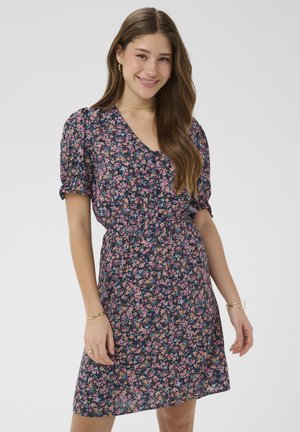 Kobieta uśmiechająca się, ubrana w granatową sukienkę w kwiaty z krótkimi bufkami na rękawach oraz złote kolczyki koła, stojąca na jednolitym tle.