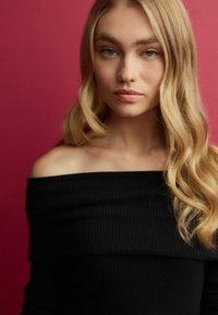 Top nero a coste con spalle scoperte, realizzato in tessuto texture, indossato con capelli biondi ondulati e sciolti, su uno sfondo rosa uniforme.