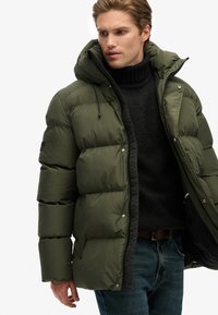 Oliven grøn dunjakke med hætte, med quiltede sektioner, lynlås og justerbare snore, båret over en sort sweater.