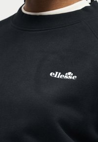 Μαύρο φούτερ με λευκό κεντημένο λογότυπο «ellesse» στο στήθος, φορεμένο πάνω από λευκό μπλουζάκι με στρογγυλή λαιμόκοψη.