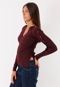 Top lungo a maniche lunghe in borgogna a coste, con scollatura a V, fessure laterali e dettagli con bottoni lungo le maniche. Abbinato a jeans blu scuro.