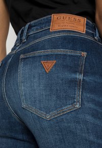 Jeans de mezclilla azul oscuro con un parche de cuero en la cintura y un logo en forma de triángulo en el bolsillo trasero. Presentan costuras en contraste.