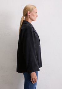 Blond naine mustas üleliises bleiseris ja sinistes teksades, seismas külg ees lihtsa heleda seina vastas.