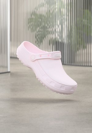 Svetloružový slip-on klož s textúrovanou podrážkou a nastaviteľným pásikom na päte, vznášajúci sa nad hladkou betónovou podlahou a kovovými vertikálnymi žalúziami.