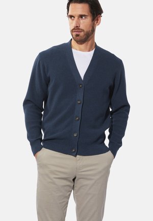 Marineblauer Cardigan aus strukturiertem Strickmaterial, mit V-Ausschnitt, sieben Knöpfen und langen Ärmeln, getragen über einem weißen T-Shirt.