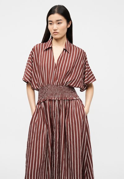 Γυναίκα με μακριά μαύρα μαλλιά που φοράει φόρεμα με καφέ και λευκές κάθετες ρίγες, με σφηκοφωλιά στη μέση και κοντά μανίκια, στέκεται μπροστά σε απλό φόντο.