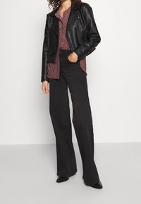 Veste de moto en cuir noir portée sur une chemise bordeaux à motifs, associée à un pantalon noir à jambes larges et des chaussures noires à bouts pointus.