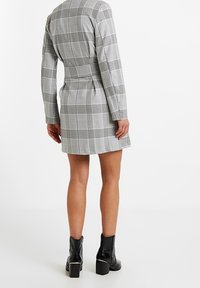 Harso kuvioinen harmaa mekko pitkillä hihoilla, vyötäröltä kireä ja takasaumassa halkio, yhdistettynä mustiin nilkkureihin, joissa on lohkokorko.