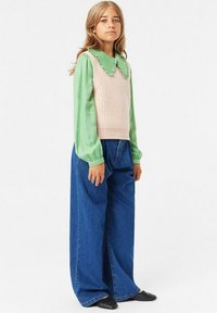 Stickad beige ärmlös tröja med ribbstruktur, lager på lager över en ljusgrön långärmad skjorta. Mörkblå vida jeans och svarta ballerinas.