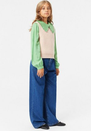 Beige gestrickter ärmelloser Pullover mit einem gerippten Muster, über einem hellgrünen Langarmshirt getragen. Dunkelblaue weite Jeans und schwarze Ballerinas.