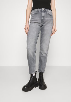 Persoon draagt vervaagde grijze high-waist jeans, zwarte enkellaarzen, witte sokken en een zwart mouwloos topje tegen een effen lichte achtergrond.