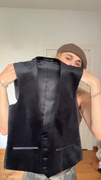Een persoon met een bruine beanie houdt een zwarte satijnen vest bij de schouders omhoog, staand binnenshuis tegen een eenvoudige muur en deur als achtergrond.