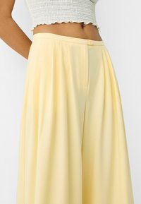 Midsection of a person wearing pastel yellow high-waisted, pleated wide-leg trousers and a white textured cropped top.