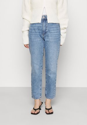 Personne portant un jean droit à taille haute bleu clair, un pull court en maille blanc et des sandales noires à talons, debout contre un mur uni.