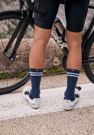 Radfahrer mit marineblauen Socken und weißen Radschuhen, der neben einem schwarzen Rennrad auf einem gepflasterten Weg steht.
