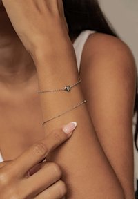 Silver chain bracelet with a small heart charm, worn on a tanned arm. Another thin silver chain bracelet is visible below.