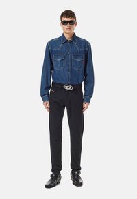 Man med mörka solglasögon, blå denimskjorta med knappar, svart bälte med silverspänne, svarta byxor och svarta läderstövlar.