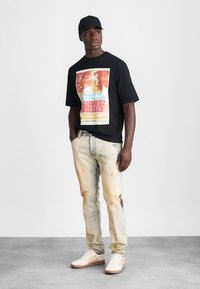 T-shirt en coton noir avec un imprimé graphique coloré, associé à un jean déchiré délavé et des baskets crème avec des rayures texturées.