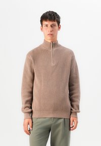 Junger Mann, der einen beigen gerippten Halbzip-Pullover und olivgrüne Hosen trägt, steht vor einem schlichten weißen Hintergrund.