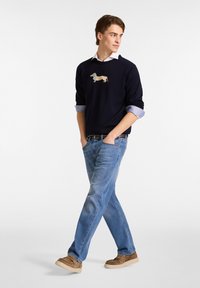 Marineblauer Pullover mit einem Dackelmotiv, kombiniert mit hellblauen Jeans und braunen Slippern. Glattes Material, lockere Passform.