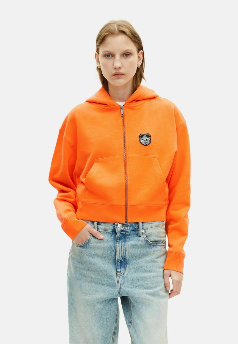 Cropped oranje sweatshirt van zachte stof met een voorkant met rits, een buidelzak en een zwart geborduurd label op de borst. Bloemendetail op het label.