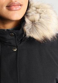 Czarny zimowy płaszcz z wysokim kołnierzem, z futrzanym wykończeniem. Widoczne metalowe zapięcia na zatrzask u szyi. Gładka tekstura materiału.