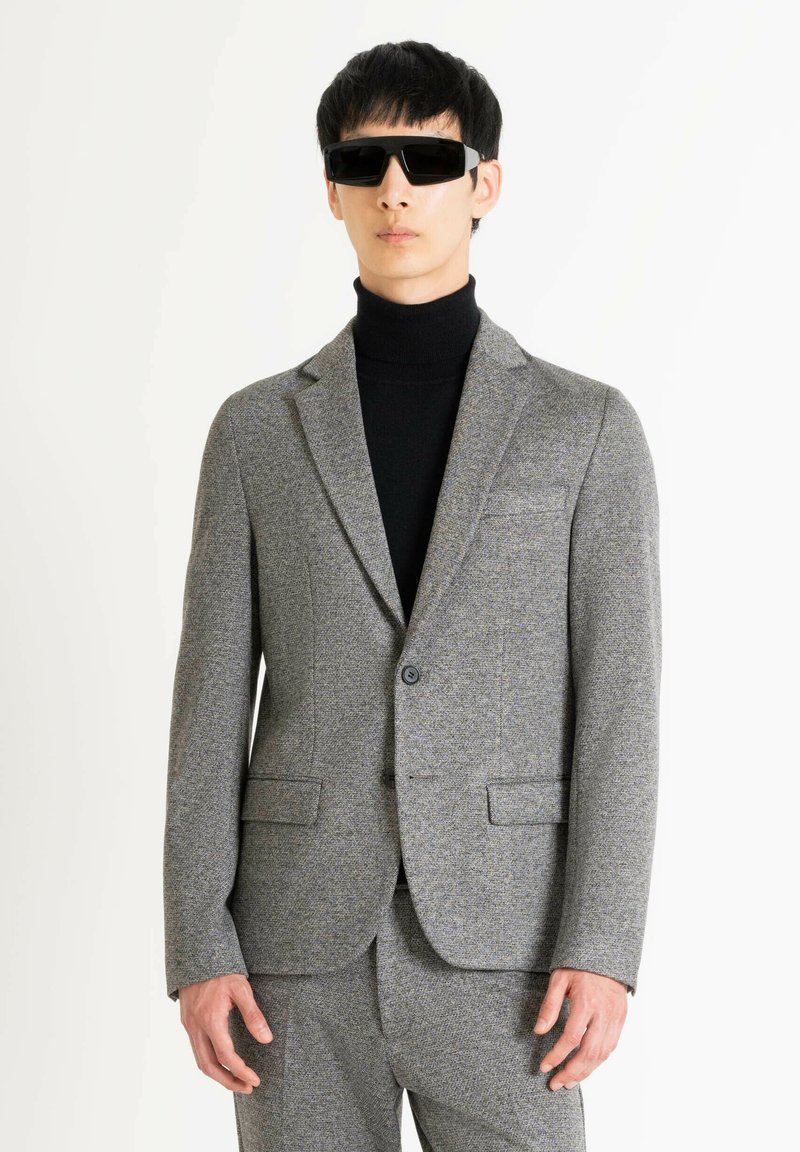 Blazer gris texturizado con dos bolsillos frontales, cierre de un solo botón y un corte entallado, combinado con un suéter de cuello alto negro y gafas de sol.