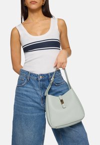 Groene leren schoudertas met een ronde vorm, gouden hardware details en een gestructureerde afwerking, vastgehouden door een persoon in high-waisted denim shorts.