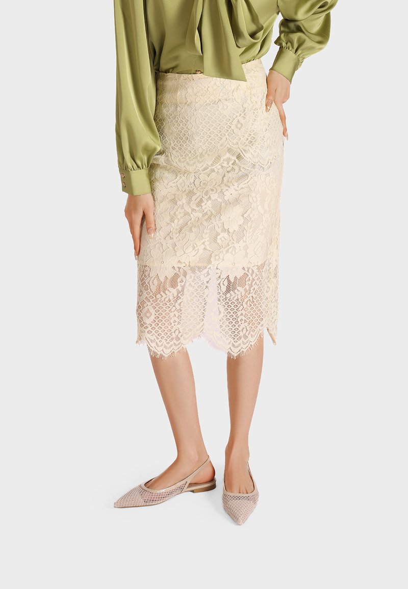 Gonna di pizzo a lunghezza ginocchio in beige chiaro con motivi floreali, orlo scallop e texture intricate, abbinata a scarpe con motivo a rete.