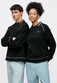 Μαύρα φούτερ με στρογγυλή λαιμόκοψη, ραφές με λευκές λεπτομέρειες και λογότυπο στο στήθος. Συνδυασμένα με ανοιχτόχρωμο μπλε τζιν.