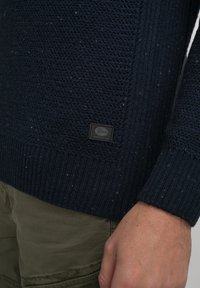 Marineblauer Strickpullover mit strukturiertem Muster, gerippten Bündchen und einem kleinen schwarzen Logopatch am Saum. Material aus Baumwollmischung.