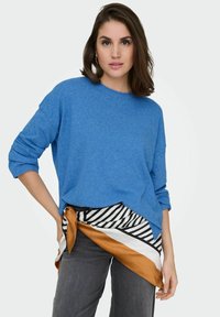 Pull en tricot bleu avec un col rond et une coupe ample, associé à une ceinture enveloppante à motifs rayés et unis autour de la taille, porté sur un pantalon gris.