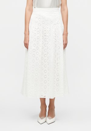 Žena nosiaca bielu midi sukňu s kvetinovým ažúrovým vzorom v kombinácii s bielymi lodičkami s čipkovým detailom.