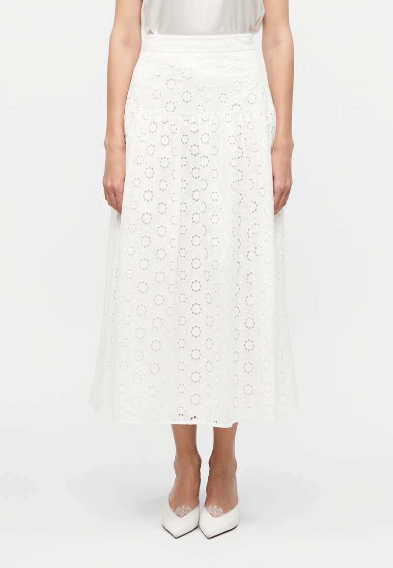 Γυναίκα που φοράει λευκή μίντι φούστα με λουλουδάτο σχέδιο από κοψίματα, συνδυασμένη με λευκές μυτερές γόβες με λεπτομέρειες δαντέλας.