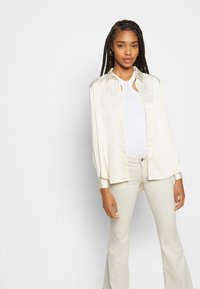Camisa de botão em cetim creme com mangas longas, combinada com uma camisola branca de alças com textura canelada e calças leves com corte flare, apresentando uma textura suave.