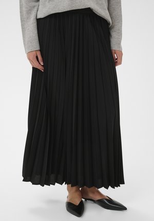 Femeie purtând o fustă midi plisată neagră, pulover gri cu mâneci lungi și pantofi negri cu vârf ascuțit, pe un fundal alb.