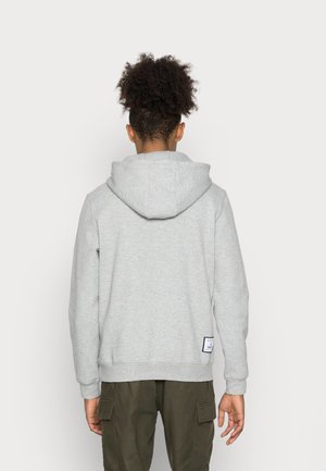 Person trägt ein hellgraues Kapuzensweatshirt und olivgrüne Cargohosen, von hinten vor einem schlichten weißen Hintergrund gezeigt.