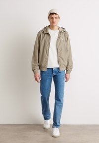 Lys khaki vindjakke med hætte, båret over en hvid t-shirt, parret med blå jeans og hvide sneakers. Modellen bærer en beige hue.