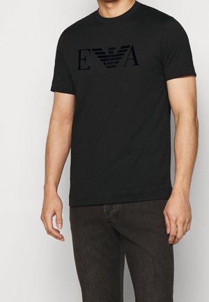 Homme portant un t-shirt noir à manches courtes avec un logo sombre sur la poitrine, accompagné d'un jean foncé, debout devant un fond uni.