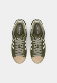 Sneakers in tessuto verde con puntali in gomma beige, caratterizzate da tre strisce bianche, occhielli rotondi e lacci grigi testurizzati.