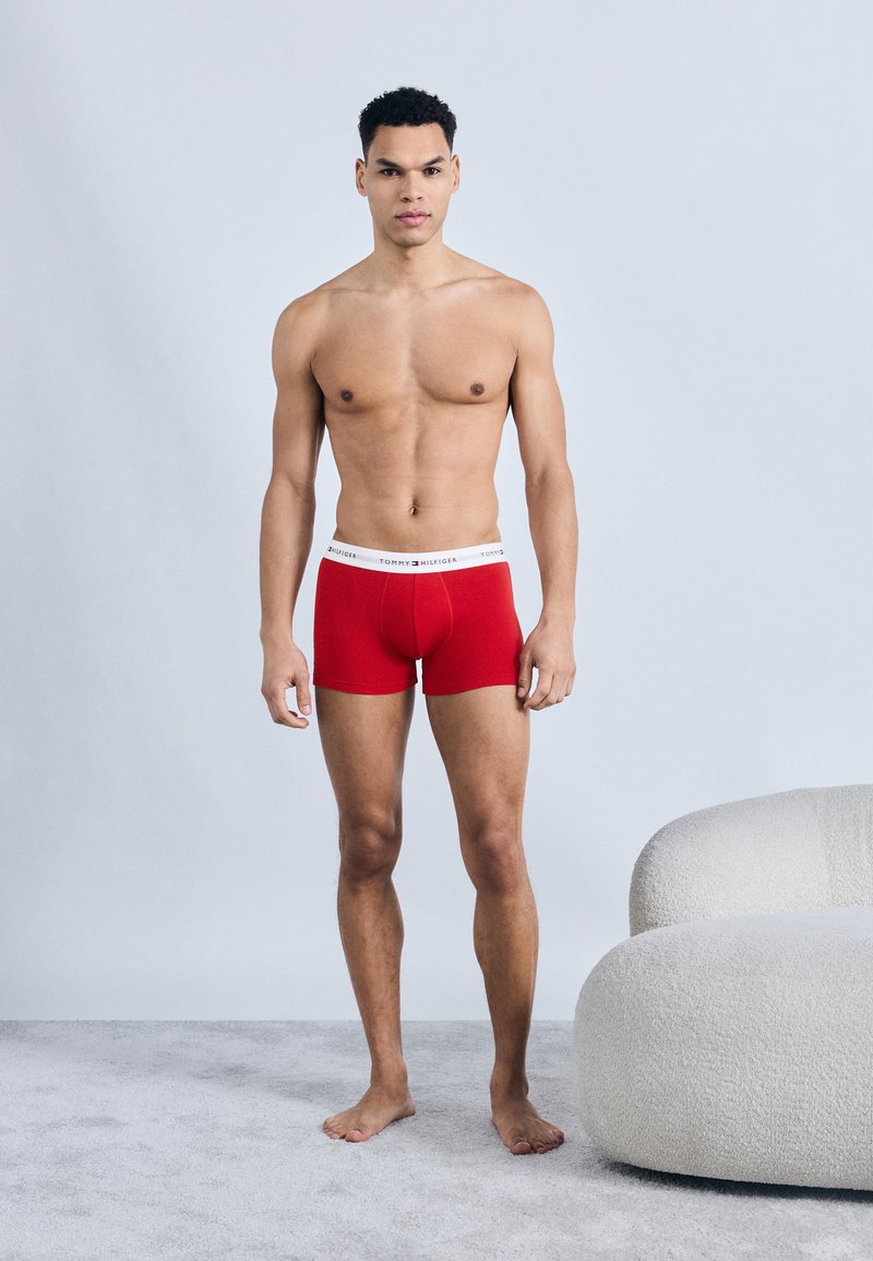Junger Mann steht barfuß auf einem Teppich und trägt rote Boxer-Shorts mit weißem Tommy Hilfiger-Bund, neben einem gebogenen weißen Sofa.