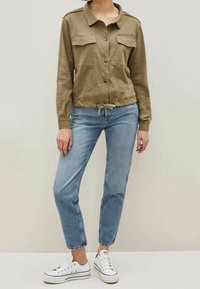 Chemise olive avec deux poches poitrine, fermeture à bouton et ourlet à cordon de serrage, associée à un jean bleu clair et des baskets blanches.