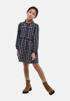 Jeune fille debout avec une jambe tendue, portant une robe à carreaux sombre avec une broderie florale et des bottines marron avec des chaussettes.