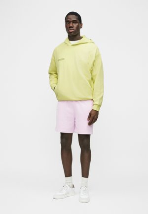 Man draagt een pastelgele hoodie, lichtroze korte broek, witte sokken en witte sneakers, staand tegen een effen witte achtergrond.