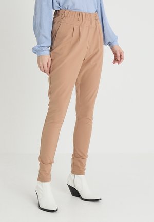 Modelka nosi beżowe spodnie z wysokim stanem i zwężane nogawki z elastyczną talią, jasnoniebieską bluzkę z długim rękawem oraz białe botki z ostrym noskiem.